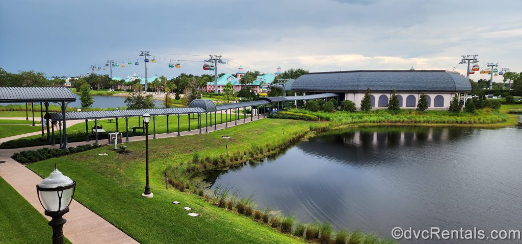 Disney’s Riviera Resort Skyliner Hub. Colorful gondolas are going to and from the hub. There is a pond nearby and a walkway going up to the hub. The sky is dark and cloudy