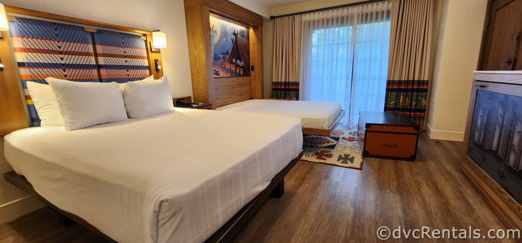 Studio at Disney’s Boulder Ridge Villas. A queen-sized bed with white linens and a pull-down bed sit in the room. A dark wood chest sits in front of the couch. The room is bright yet has rustic touches.