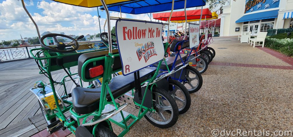 A line of colorful surrey bikes can be seen sitting on the Boardwalk. The back of the first surrey bike has a sign that reads “Follow me to Disney’s Boardwalk.”