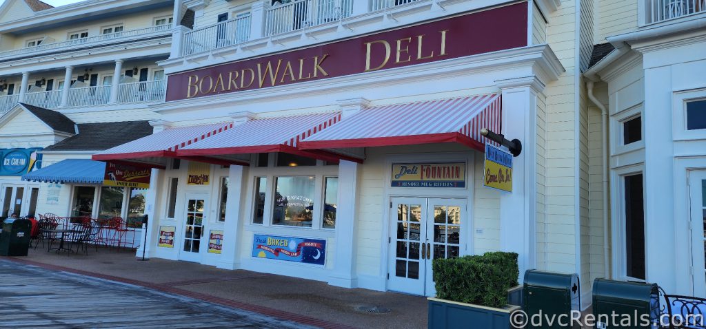 Boardwalk Deli Storefront. A red Sign with Gold Lettering spells out Boardwalk Deli. Underneath, red and white awnings hang above glass doors and windows.