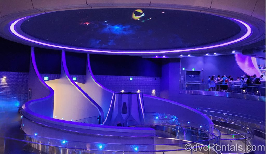 Inside Queue for Guardians of the Galaxy. The lighting is dark blue and purple, the room is very sleek and futuristic, and there is a moving photo of space projected on the ceiling.