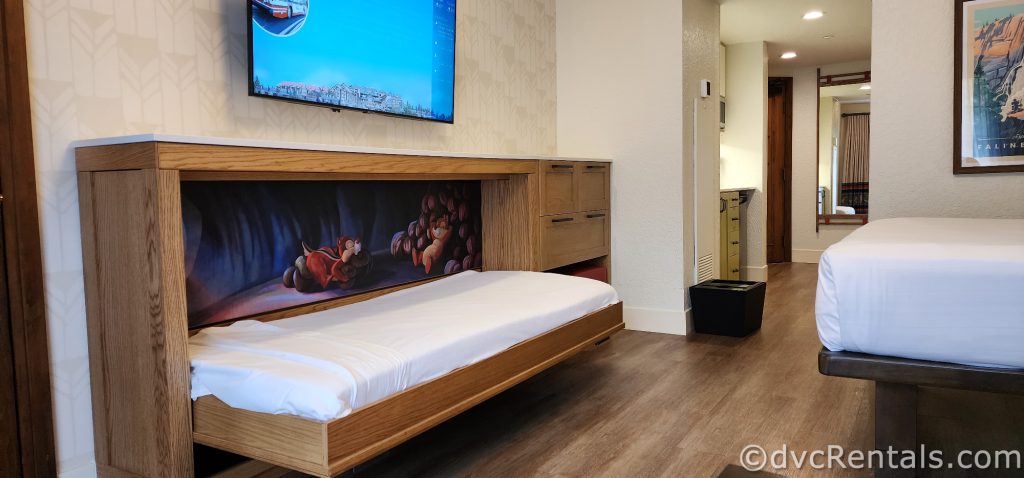 A down bunk is pulled out from a wooden console. Painted behind the bed is a mural of Chip and Dale sleeping on acorns in a cave.