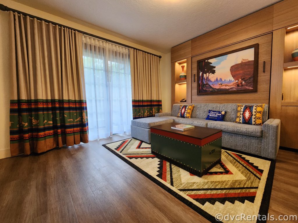 Living Room in the Boulder Ridge One Bedroom. There is a large gray couch and stool with a dark green and red chest sitting in front of it. A tribal pattern carpet sits on the dark hardwood floors, and behind the couch hangs a mural of Mickey Mouse and Pluto going hunting.