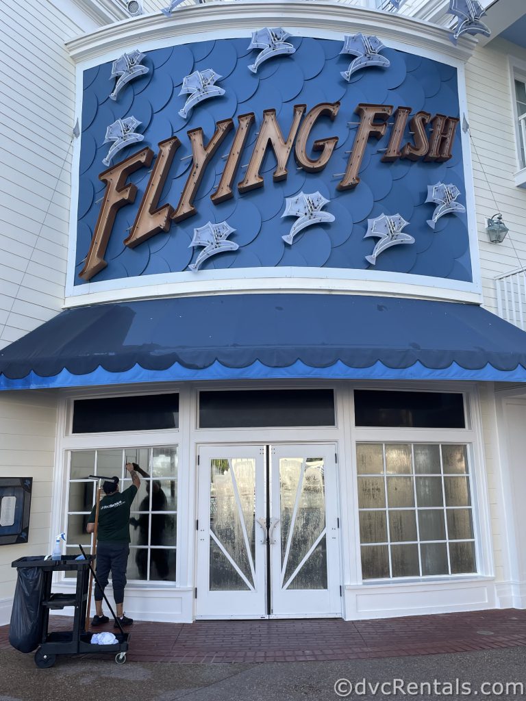 Flying Fish restaurant. The building is white with a large blue and gold sign. There is a window washer cleaning the front windows.