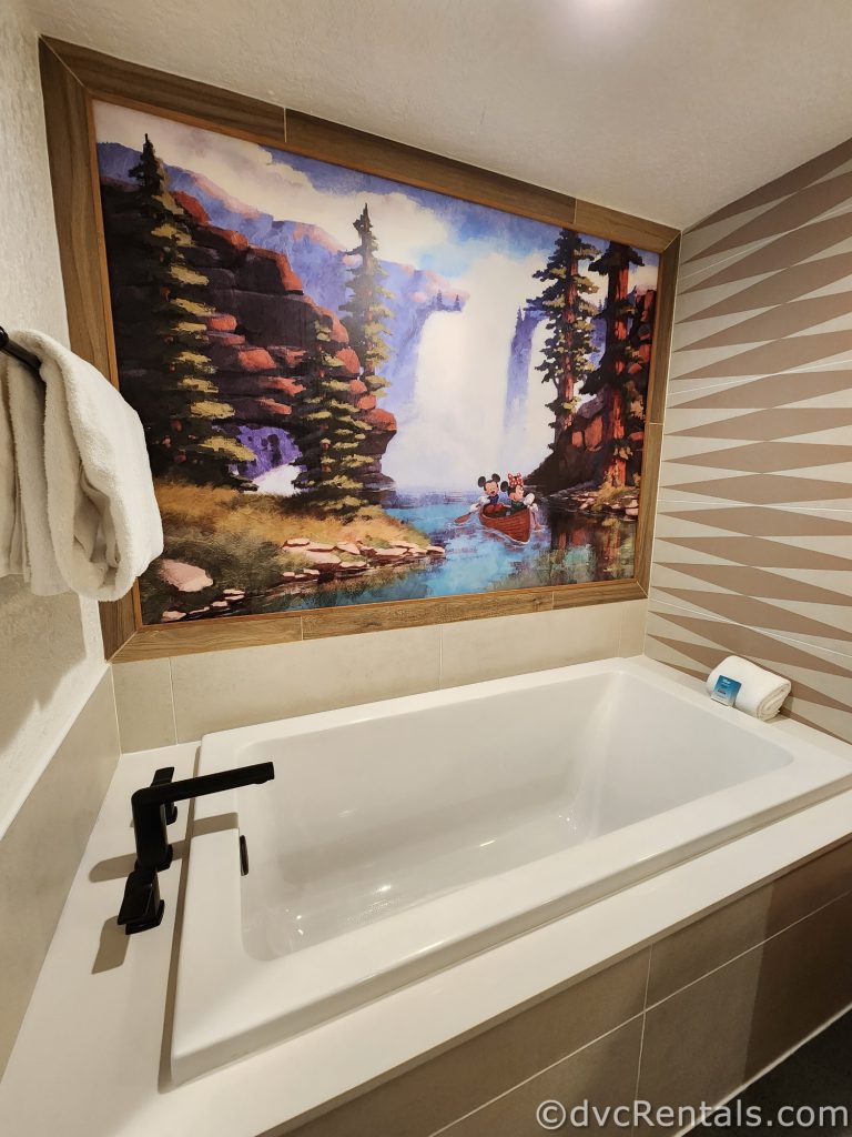 A large white porcelain tub. Painted behind the tub is a mural of Mickey Mouse and Minnie Mouse kayaking down a river with a forest on either side.