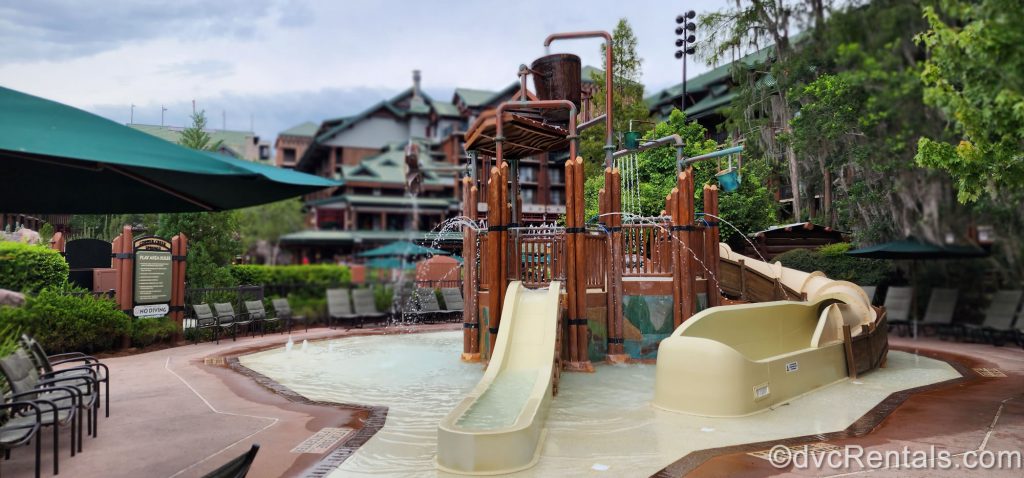 Kids waterplay area at Copper Creek Springs Pool at Disney’s Wilderness Lodge. The faux-wood waterplay jungle gym includes a bucket pouring out water at the top and two yellow slides. There are sprinklers of water shooting out from all sides of the Jungle Gym.