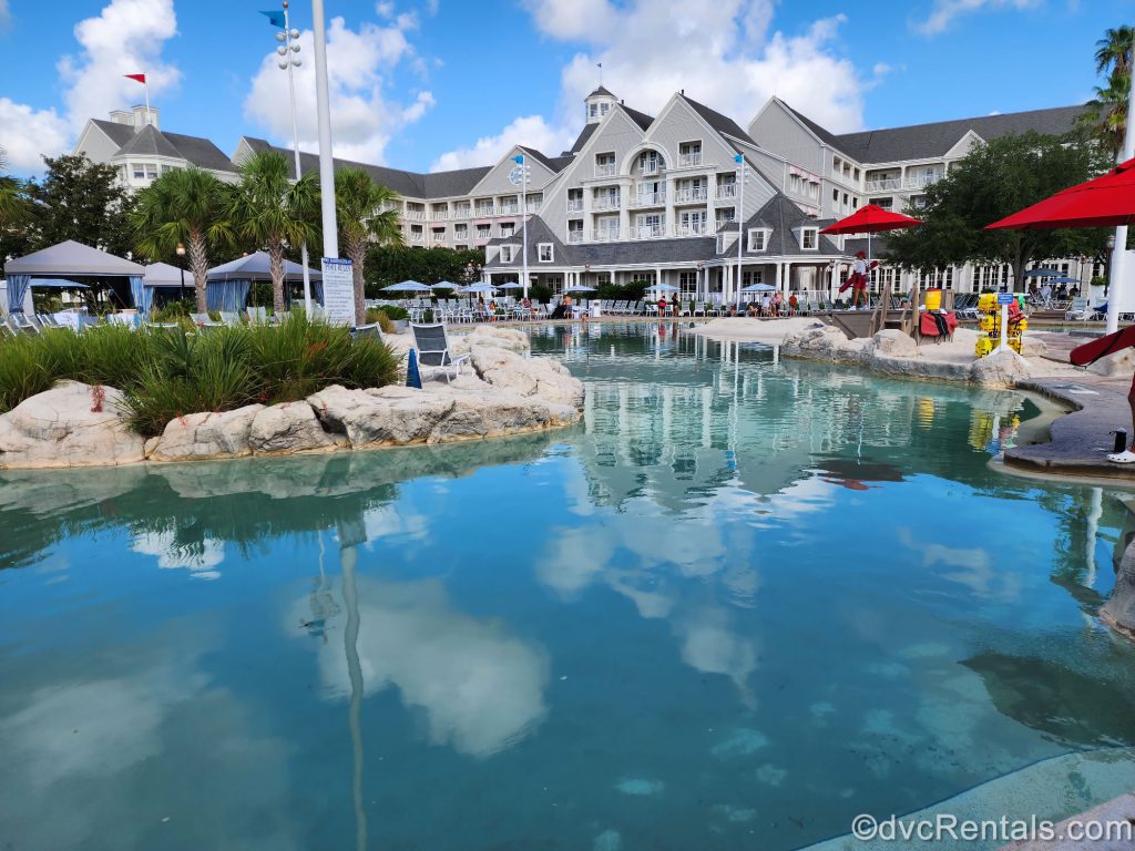 Stormalong Bay at Disney’s Beach Club Resort. There are gray rocks on either side of the pool and some red umbrellas around the pool. You can see Disney’s Yacht Club Resort in the distance.