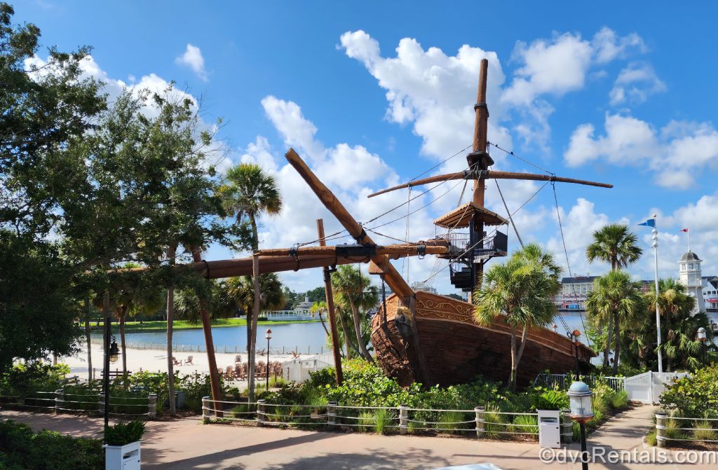 Pirate-Ship Waterslide at Stormalong Bay. The ship appears to be made of wood, and the slide runs in the air above the sidewalk. There are bushes and trees surrounding the ship. 