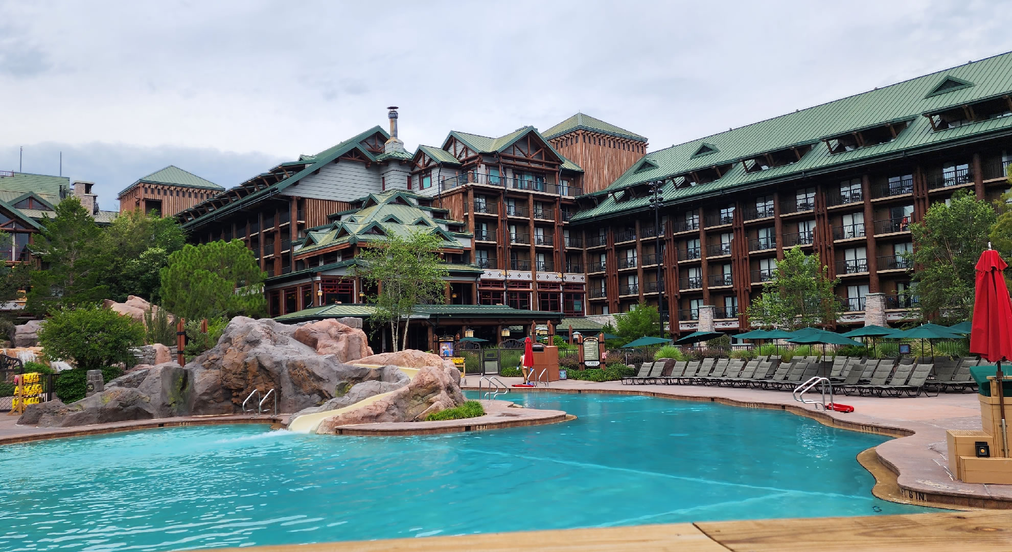 Copper Creek Springs Pool at Disney’s Wilderness Lodge. The log cabin-esque building with green accents can be seen in the background of the photo. A waterslide surrounded by fake rocks flows into the pool.