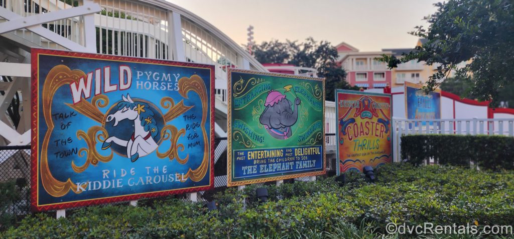Carnival-Themed Painted Billboards at Disney’s Boardwalk Villas. The three billboards stand within green bushes. The first billboard includes a picture of a white horse and says, “Wild Pygym Horses, Ride the Carousel.” The second billboard includes a picture of an elephant and reads, “Entertaining and Delightful, The Elephant Family.” The third billboard reads “Keister Coaster Coaster Thrills.”