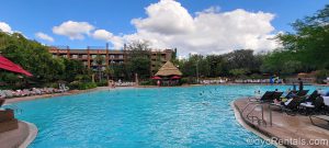 Uzima Springs Pool at Disney’s Animal Kingdom Lodge. Several people are swimming in the blue water of the pool. The resort can be seen in the background, and beige lounger chairs surround the pool deck.