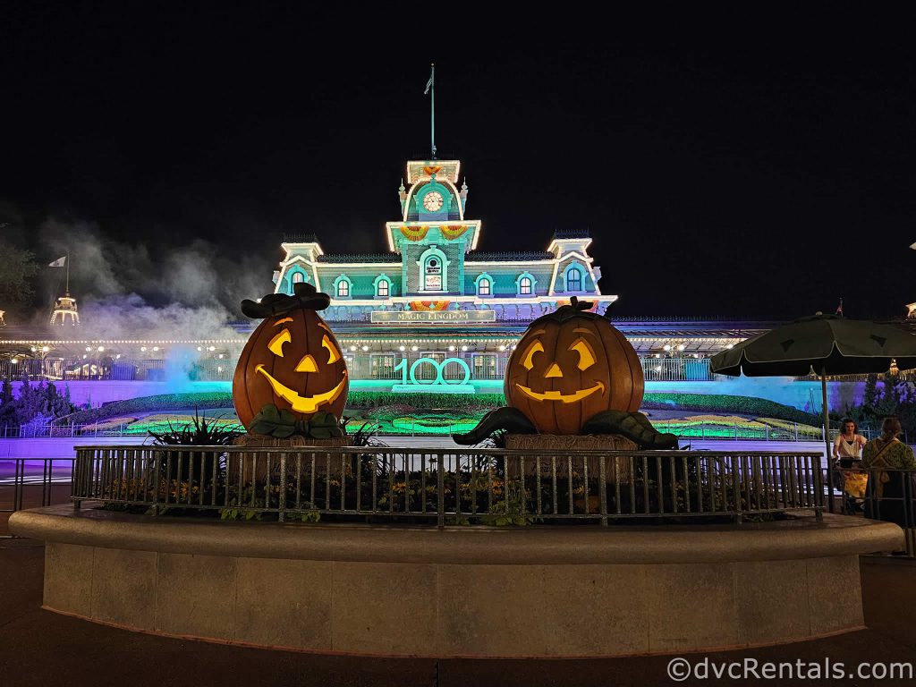 Two large pumpkins sitting in a garden, with the Magic Kingdom Railway Station illuminated in the background.