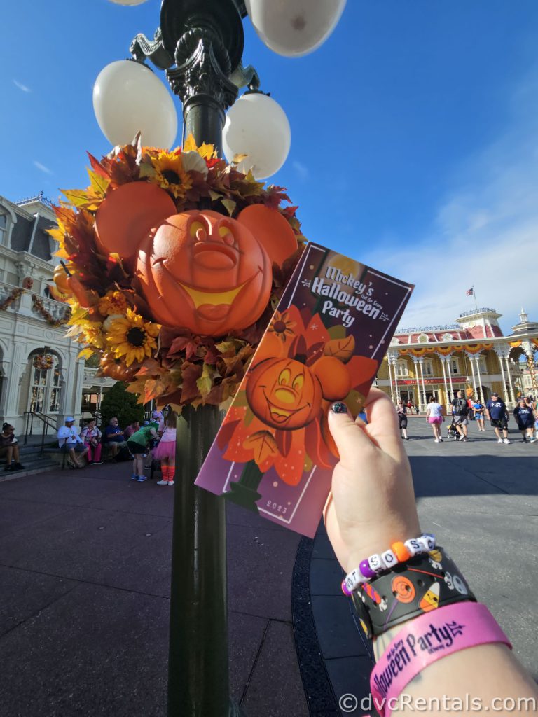 Mickey Mouse wreath hanging on a lamp post. A hand is holding up the Halloween Party pamphlet in front of the lamp post.