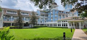 Front of Disney's Beach Club Resort. Bright blue building with white trim and porch. There is grass in front of the building and trees overhead.