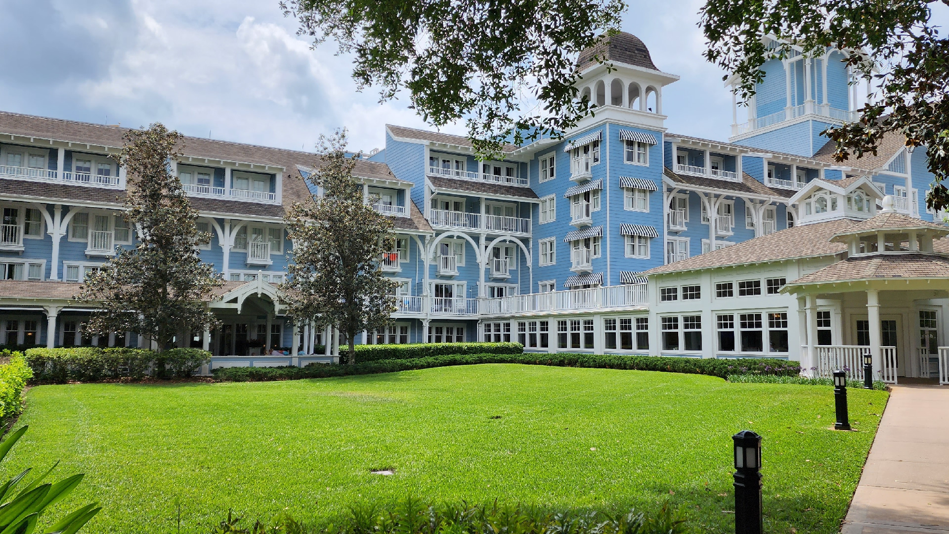 Front of Disney's Beach Club Resort. Bright blue building with white trim and porch. There is grass in front of the building and trees overhead.
