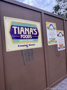 A sign that reads "Tiana's Foods Coming Soon" sits on a brown Construction Wall with a couple of other signs blurred in the background.