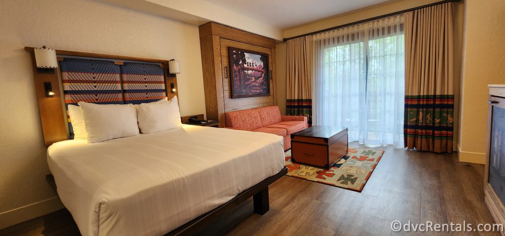Studio at Disney's Boulder Ridge Villas. A queen-sized bed with white linens and a red patterned couch sit in the room. A dark wood chest sits in front of the couch. The room is bright yet has rustic touches.