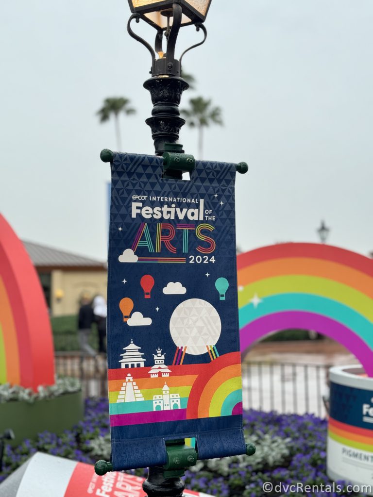 Festival of the Arts Sign hanging from a green pole with out-of-focus rainbow statues in the background.