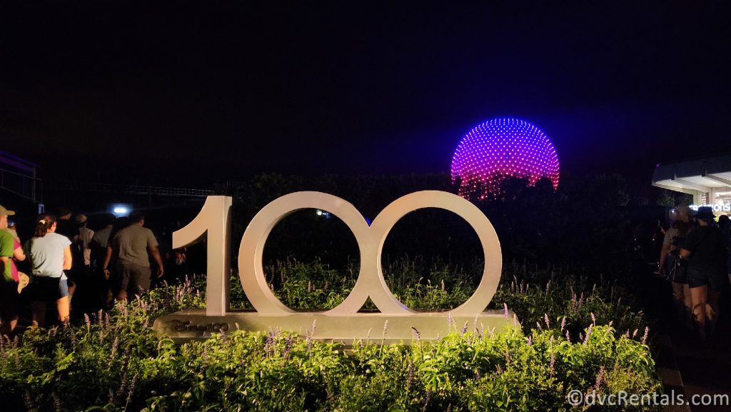 Disney 100 Sign at night with the illuminated Spaceship Earth in the background.