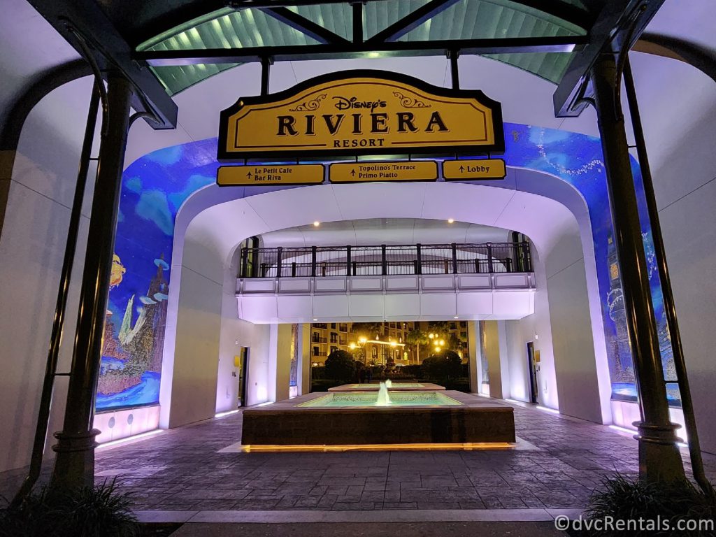 Entrance to Disney's Riviera Resort from the Skyliner hub at night. The murals on either side of the walls are lit up with a fountain between them.