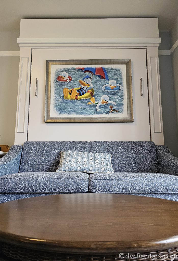Painting of Donald Duck, Huey, Dewey, and Louie above a blue couch. There is a throw pillow on the couch and a dark wood coffee table in front of the couch.