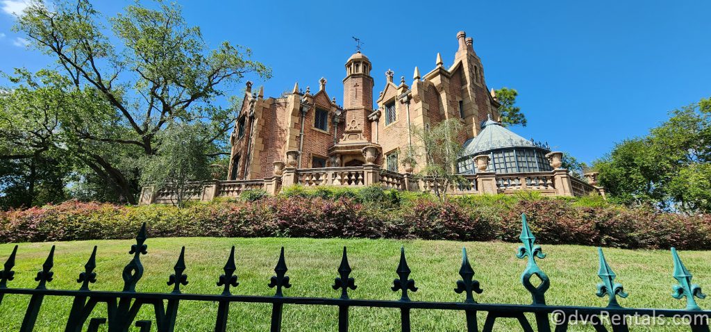 Exterior of the Haunted Mansion in Magic Kingdom. The brown building is at the top of a grassy hill, and a rod-iron fence is at the front of the photo.