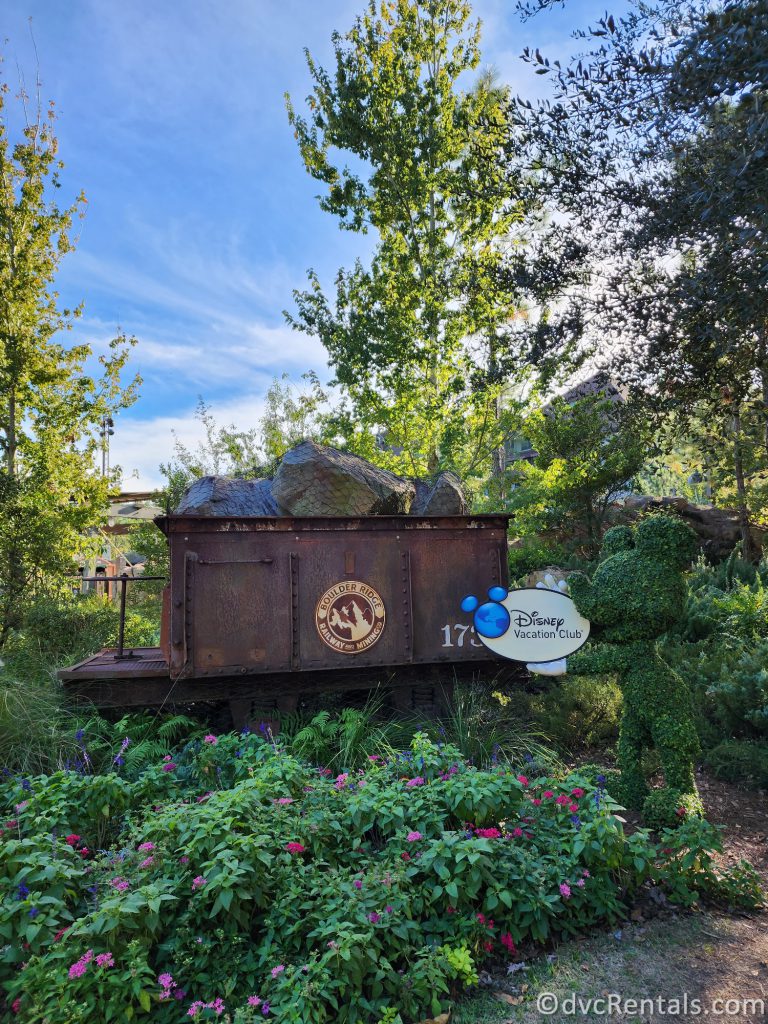 Grass Topiary of Mickey Mouse holding a sign that reads "Disney Vacation Club" next to a brass dumpster holding boulders that reads "Boulder Ridge."