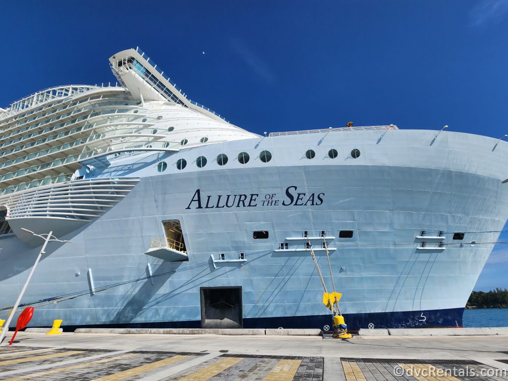 Front of Royal Caribbean's Allure of the Seas ship docked at port. The white ship has the words "Allure of the Seas" painted in blue on the side.