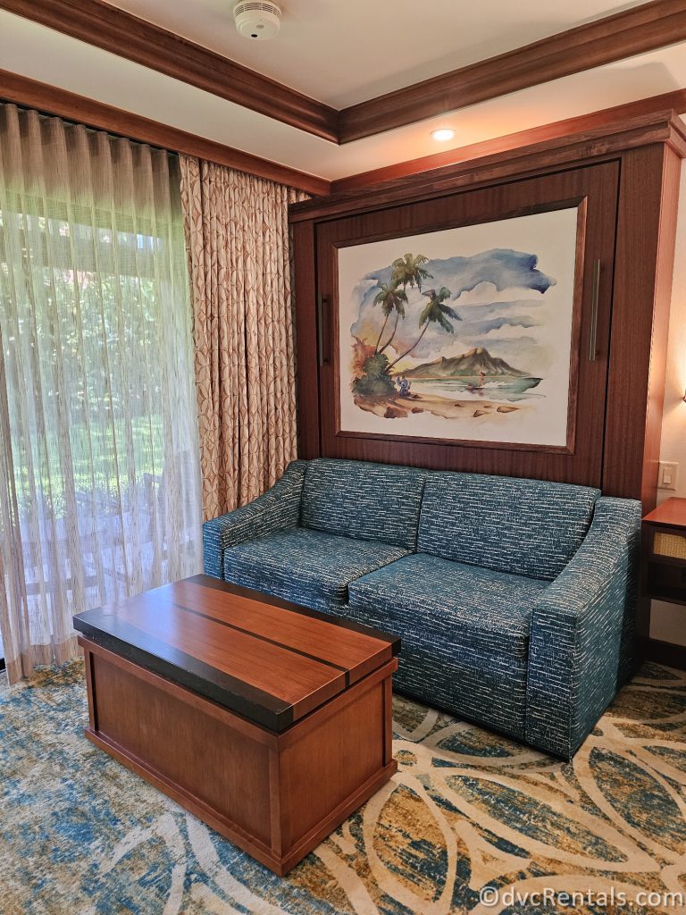 Blue Fabric couch in the studio at Disney's Polynesian Villas. There is a wooden coffee table sitting in front of the couch and a print of Stitch on the beach playing a ukulele above the couch.