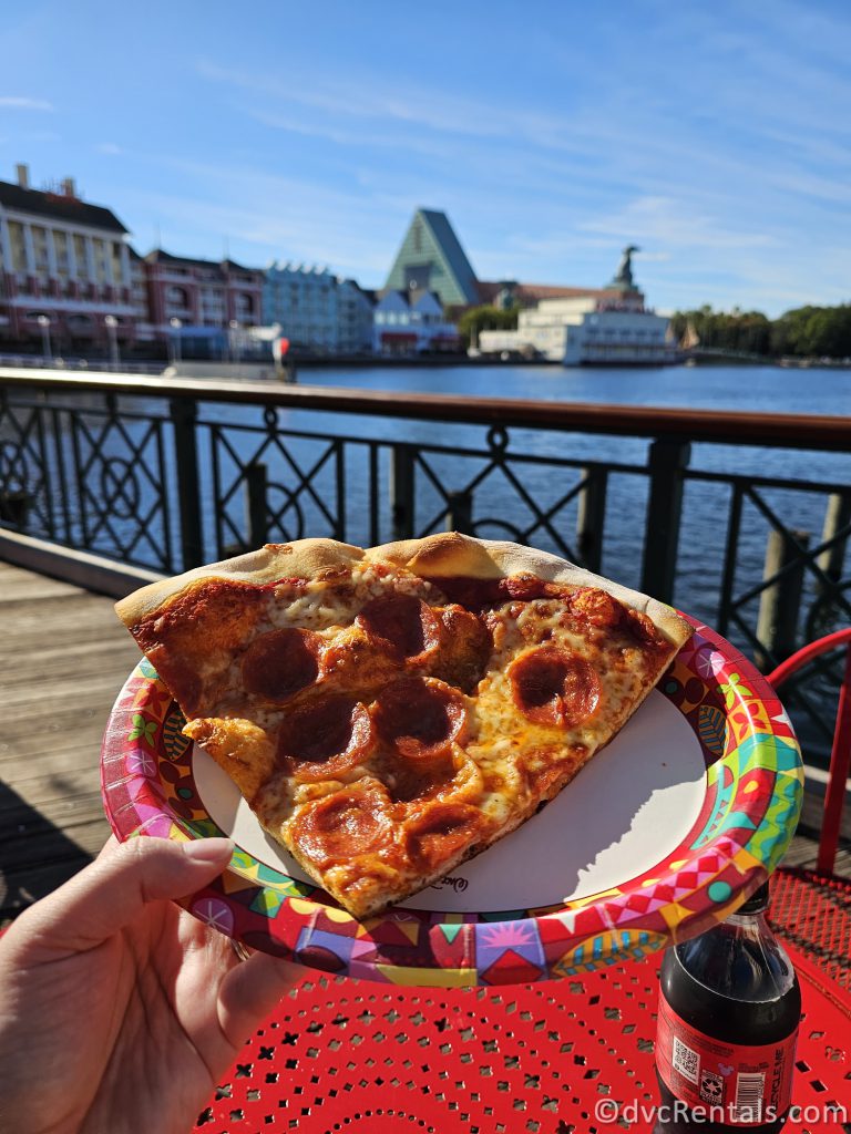 Slice of pepperoni pizza on a paper plate with the water from Crescent Lake and the Boardwalk in the background.