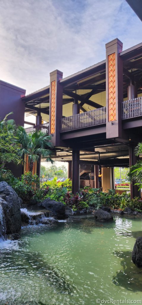 Exterior of Great Ceremonial House. There is a pond surrounded by tropical flora in front of the dark wood building.