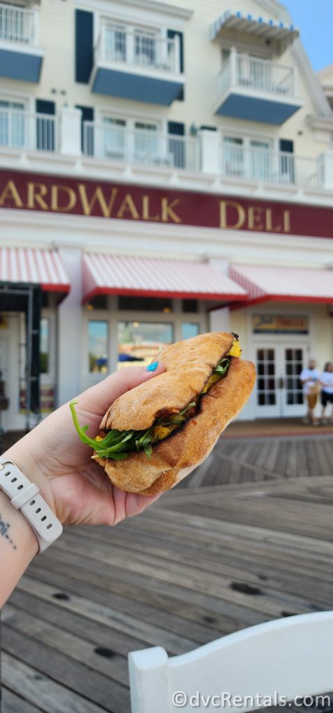 Hand holding a sandwich in the air with the sign of the Boardwalk Deli blurred in the back of the photo.