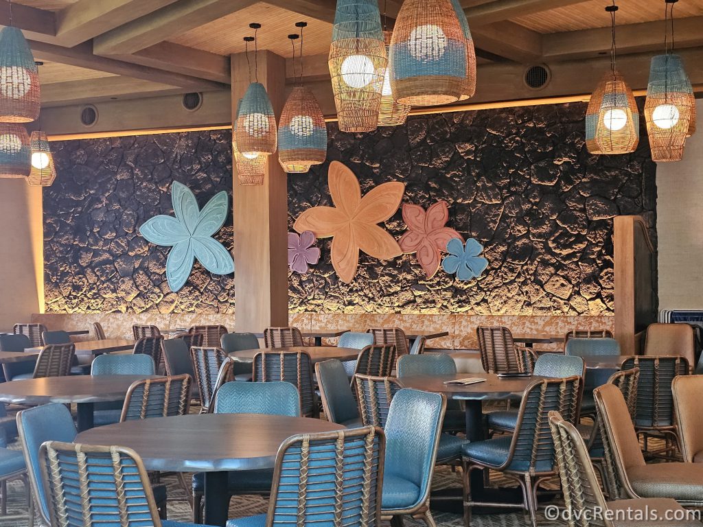 Interior of Kona Cafe. There are large wooden flowers hanging on the walls and tables and chairs throughout the restaurant.
