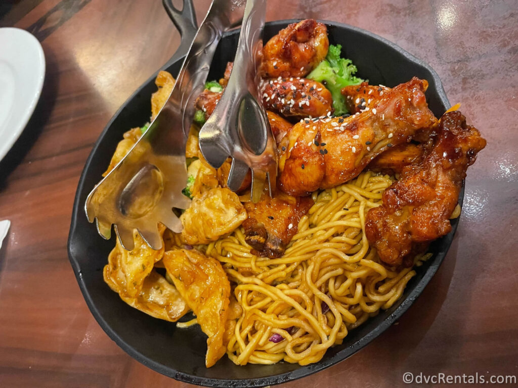 'Ohana appetizer platter featuring noodles, chicken wings, vegetables, and potstickers sitting in a cast iron pan.