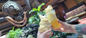 One cup of dole whip and one dole whip float being held up in front of the Tiki statue at Disney's Polynesian Villas.