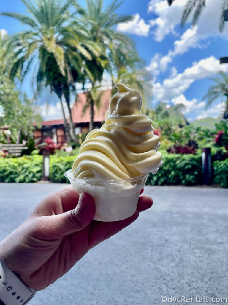 Pineapple Dole Whip in a clear plastic cup being held up in front of the building at Disney's Polynesian Villas.