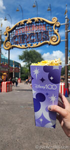 Purple carton of popcorn being held up in front of the Storybook Circus sign in Magic Kingdom.