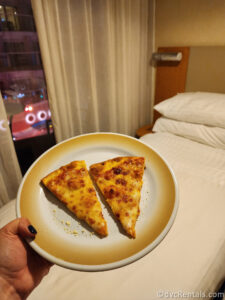 Two slices of cheese pizza sitting on a plate. The plate is being held over a bed in a stateroom.