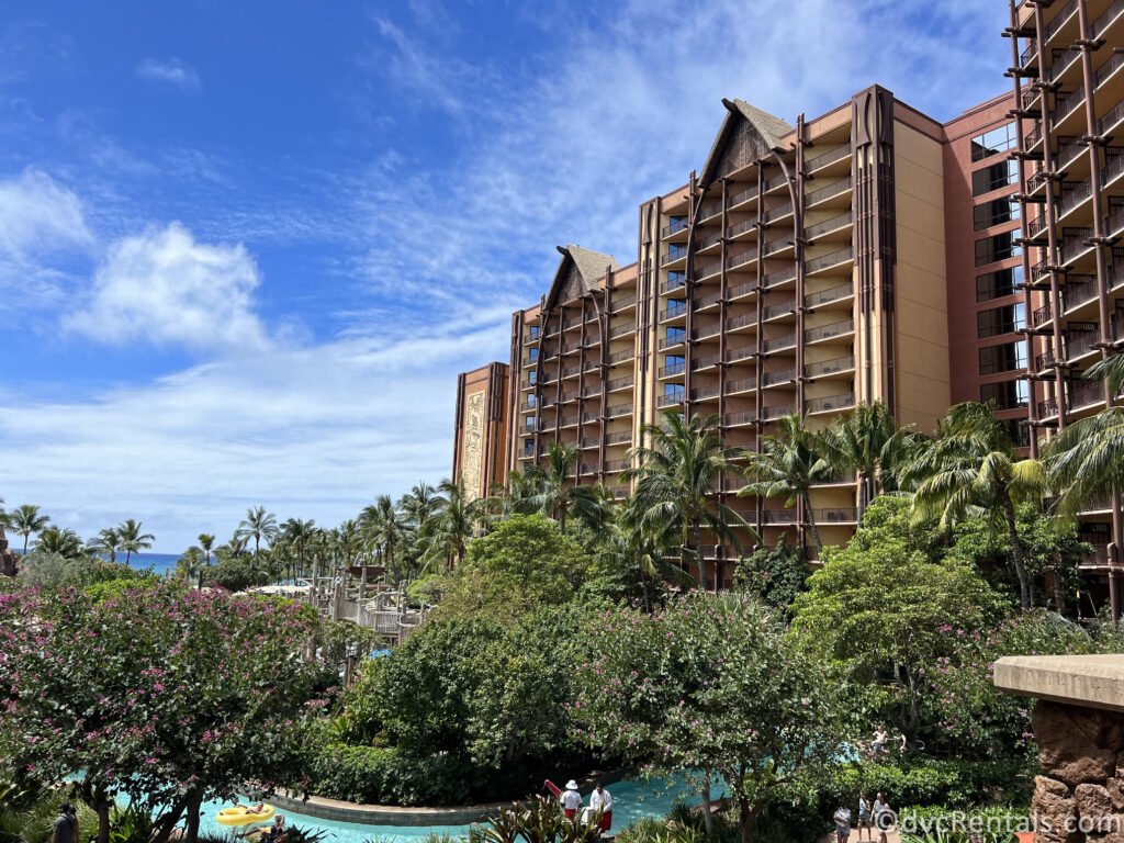 Exterior of Aulani, with green trees and a lazy river in front of the building.