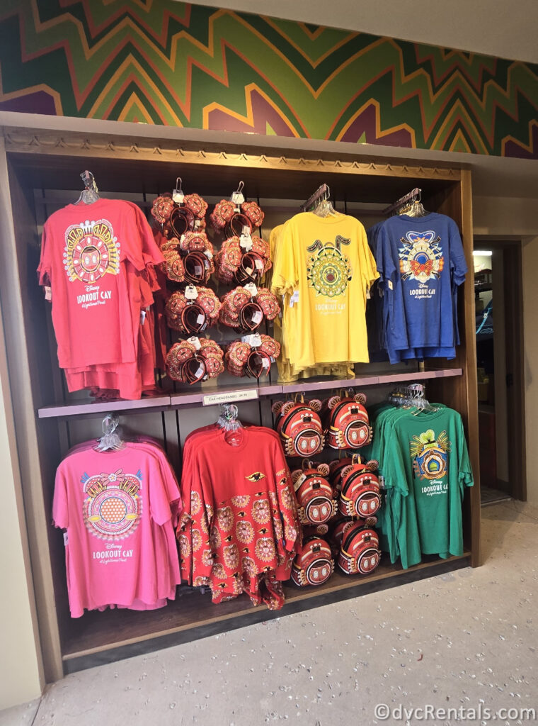 Colorful Lookout Cay at Lighthouse Point t-shirts, Mickey ears, and lounge fly backpacks hanging on a wall.