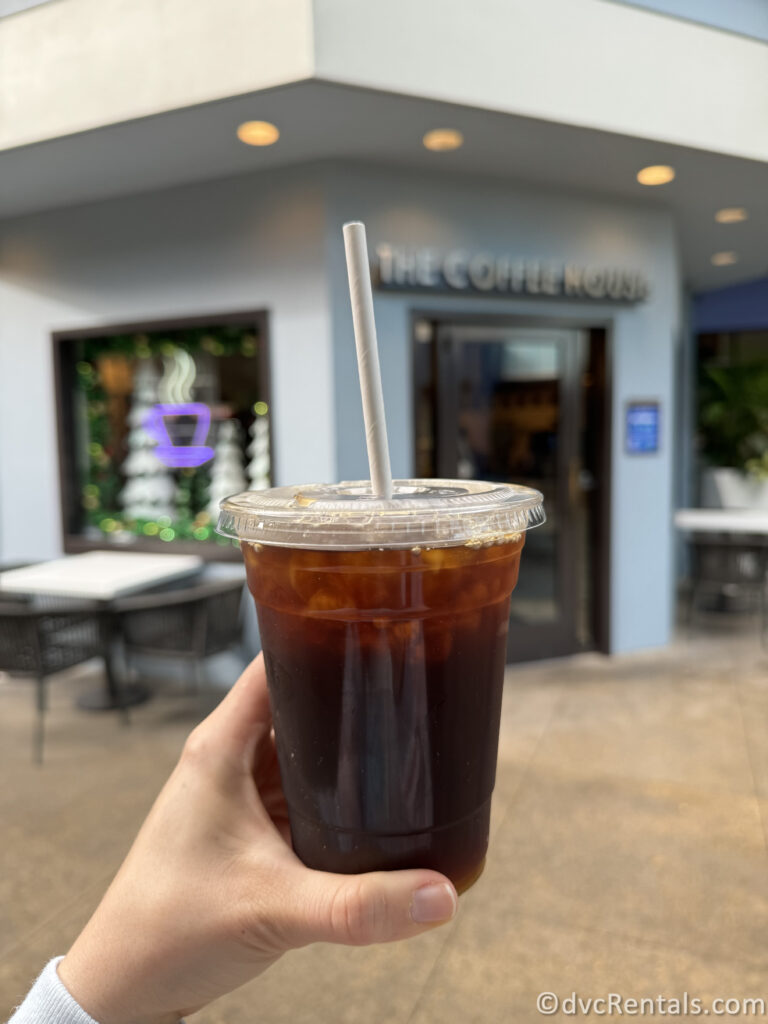 Black Iced Coffee in a plastic cup being held in front of the entrance to The Coffee House, which is blurred in the background of the photo.