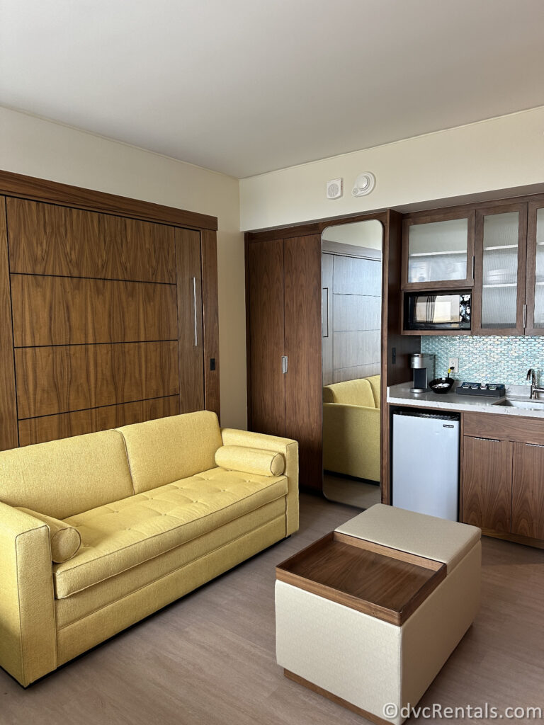 Living area inside the Studio. There is a yellow couch and a kitchenette that is covered in dark wood.