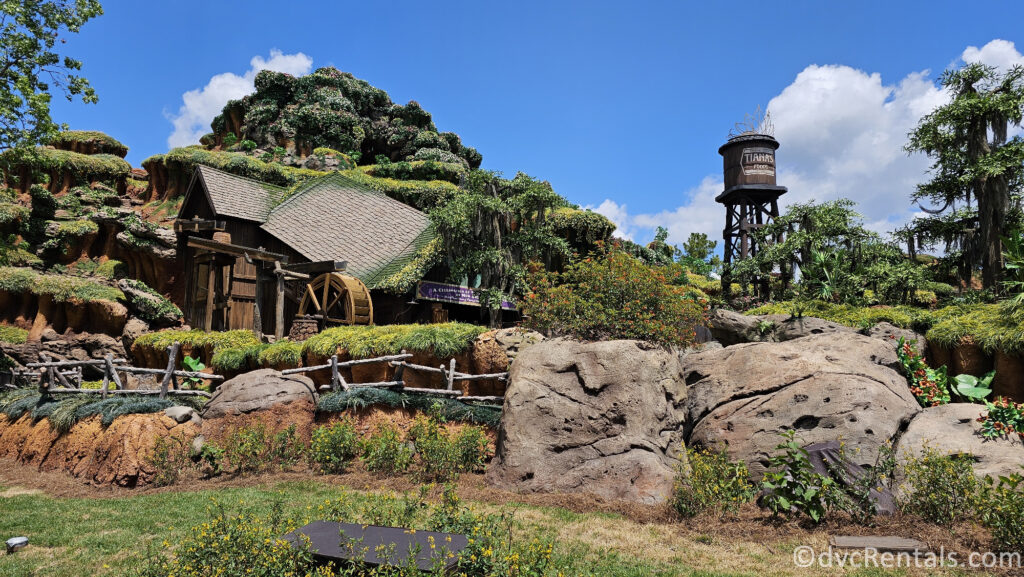 Exterior of Tiana's Bayou Adventure. Greenery and rocks cover a large hill, and there is a brown water tower in the background.