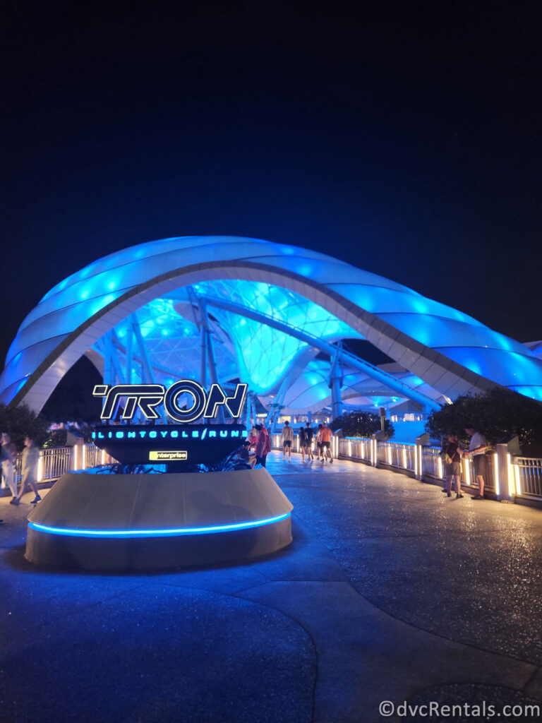 Entrance to Tron: Lightcycle/Run. The entrance sign and the ride in the background are both glowing blue in the night sky.