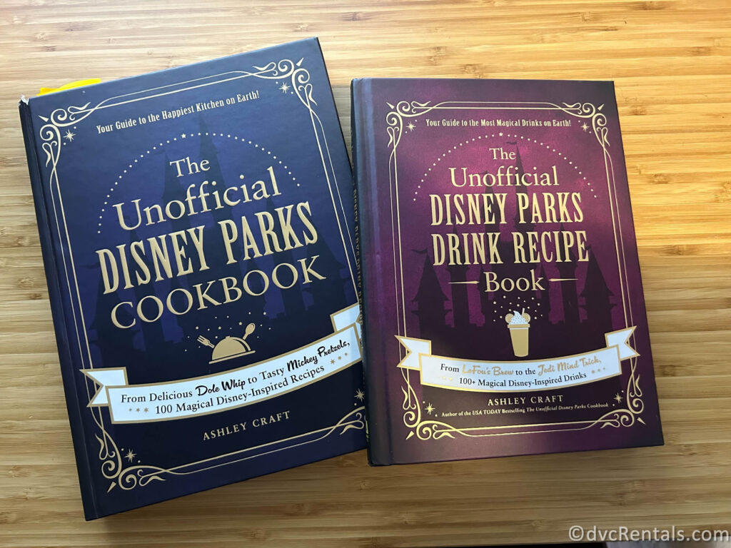 Two cookbooks sit next to each other on a wooden table. One reads "The Unofficial Disney Parks Cookbook" and the other reads "The Unofficial Disney Parks Drink Recipe Book."