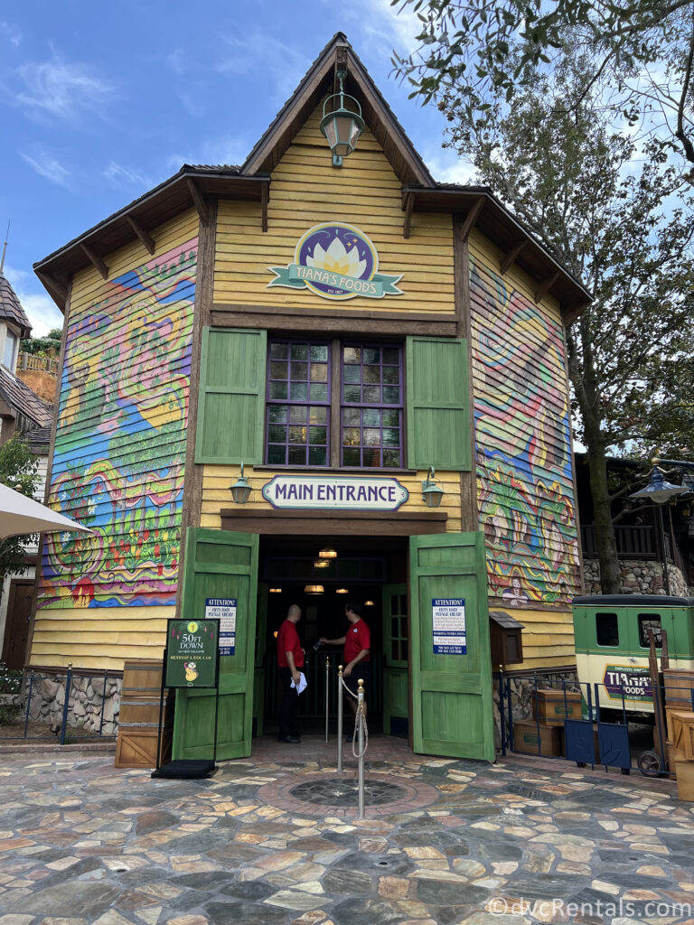 Entrance into Tiana's Bayour Adventure. Bright yellow wood building featuring murals on the side, and green doors open to welcome everyone into the queue.