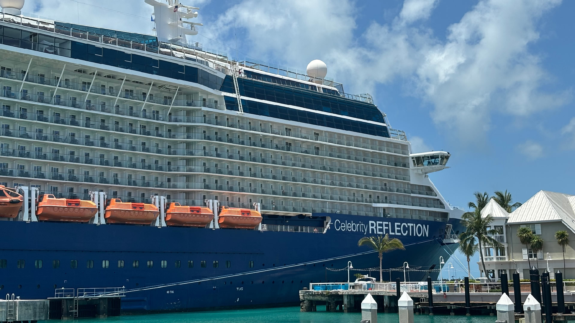 The Cruise Ship "Celebrity Reflection" docked at a harbor with several lifeboats attached along its side. The ship is blue and white, with multiple balconies for guest cabins.