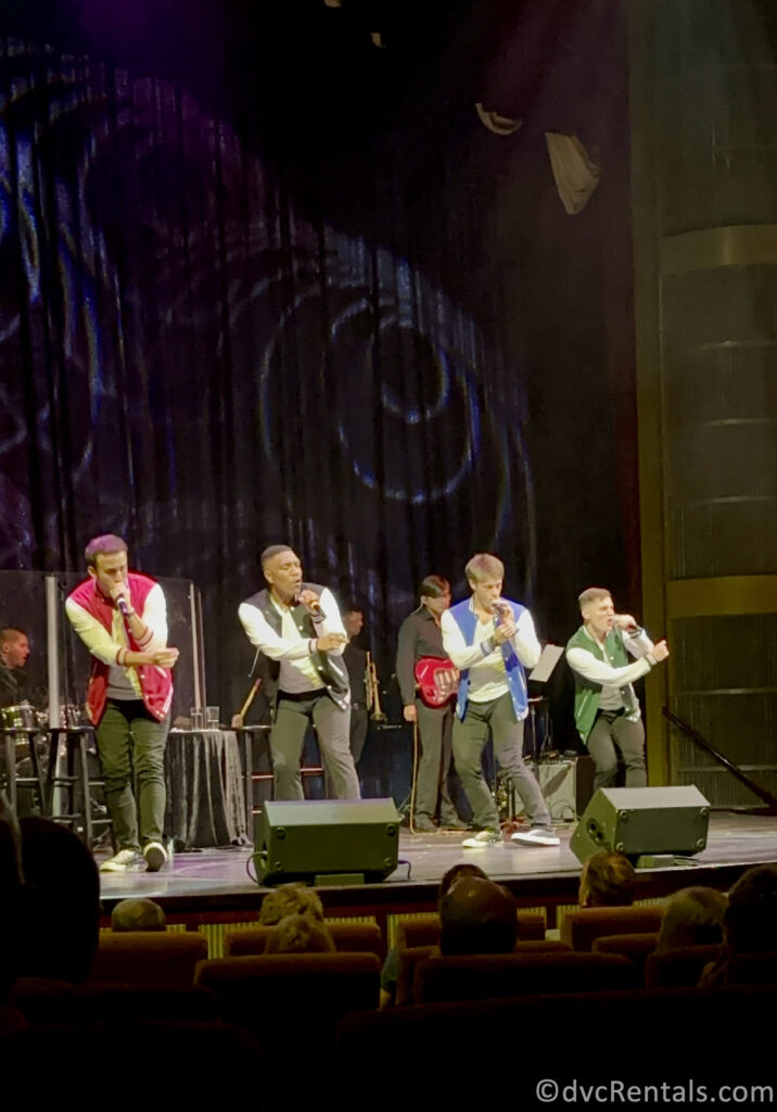 Four men dancing on a stage in different colored shirts singing into microphones.