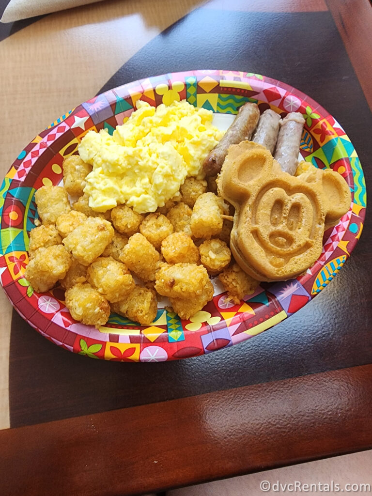 Classic Breakfast combo sitting on a paper-to-go plate. There are scrambled eggs, sausage links, tater tots, and Mickey Waffles sitting on the plate.