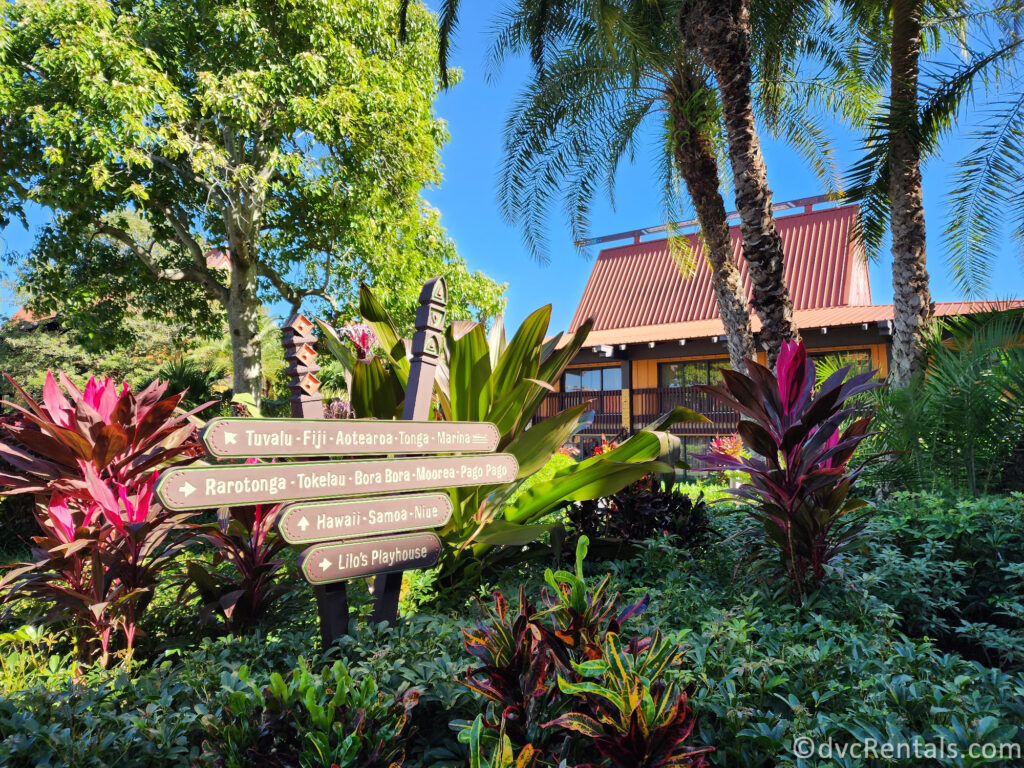 Signpost in front of a Polynesian-style building at Disney's Polynesian Village Resort. The building has a thatched roof and colorful accents, and there are lush tropical plants surrounding it.